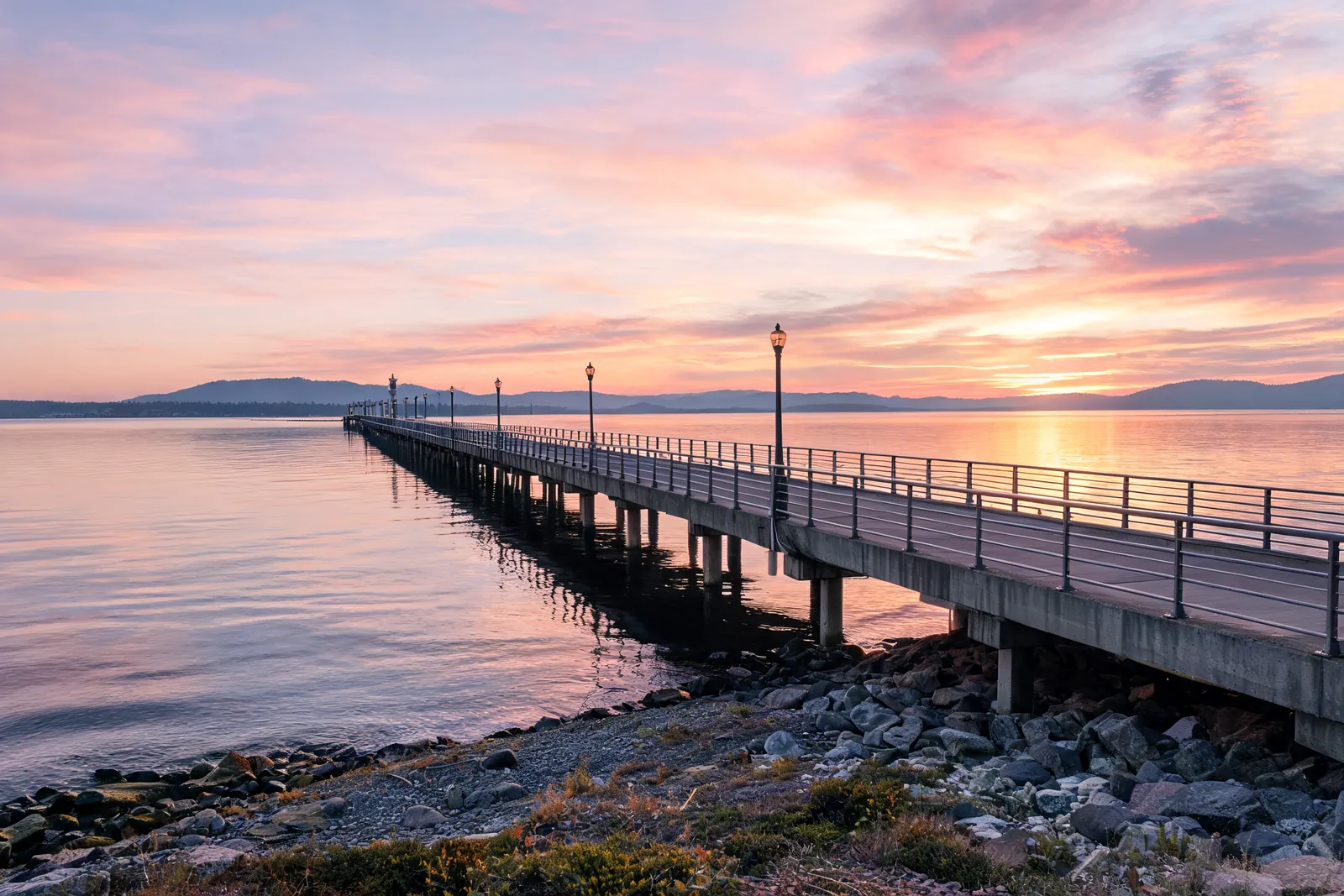 A long public pier with lamp posts stretching over calm water under a pink and orange sunset